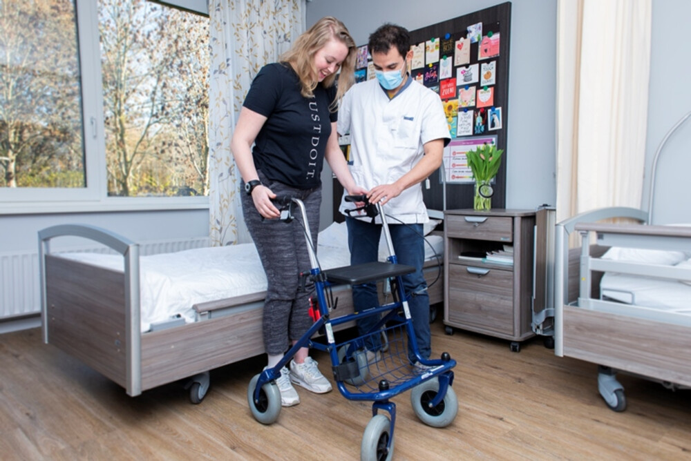Romy wordt geholpen door haar verpleegkundige met lopen achter een rollator.